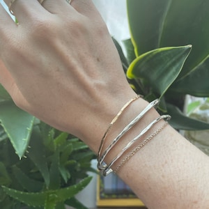 Pode incluir: Um conjunto de quatro pulseiras de prata e ouro no pulso de uma pessoa. As pulseiras estão empilhadas umas sobre as outras e são feitas de metal martelado.