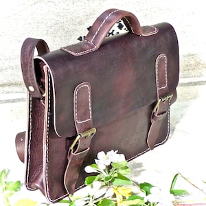 Peut inclure: Un sac à main en cuir marron avec une poignée supérieure et une bandoulière. Le sac a deux boucles sur le devant et des coutures blanches.