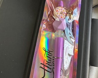 Op de afbeelding: Een paarse pen met een zilveren punt en decoratieve bedels, verpakt in een holografische zak. De bedels omvatten een donut, kralen en een transparante bol. Op de zak staat "Thank you".