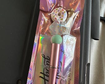 Op de afbeelding: Een roze en groene make-up kwast met een mintgroene spons tip, verpakt in een holografische roze zak. De zak bevat een donut bedel, een bliksem bedel en een gouden bedel. Een rode sticker met het nummer 1001 is zichtbaar.