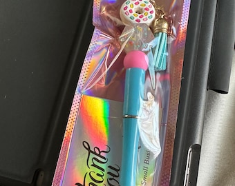 Op de afbeelding: Een blauwe pen met een roze bovenkant en een donut-bedel. De pen is verpakt in een holografische zak met de woorden "thank you" en "For Supporting My Small Busi" gedrukt op een kaart.