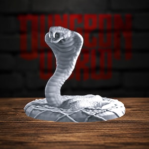 Pode incluir: Uma estatueta em miniatura de cobra cinza-claro com a boca aberta e presas visíveis, enrolada numa base circular com um padrão de rede texturizado. A cobra está numa posição de ataque ereta com o capuz aberto, exibindo escamas detalhadas.