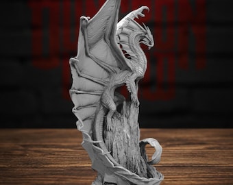 May include: A gray, unpainted dragon figurine standing on a textured rock formation. The dragon features large, detailed wings that extend upward, sharp claws, and a long tail that curls around the base of the rock. The surface is matte.