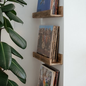May include: Three wooden shelves mounted on a white wall, each shelf holds a stack of vinyl records. The top shelf has a blue record with a person swimming on the cover. The middle shelf has a record with a group of people on the cover. The bottom shelf has a record with a colourful abstract design on the cover.