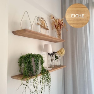 Könnte beinhalten: Zwei schwebende Holzregale an einer weißen Wand, die dekorative Gegenstände präsentieren. Das obere Regal enthält Glasvasen und Trockenblumen. Das untere Regal zeigt eine Topfpflanze, eine Lampe und Glasvasen. Das Wort "EICHE" ist in einem Kreis sichtbar.