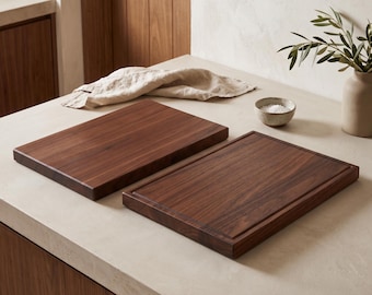 May include: Two rectangular dark brown wooden chopping boards with smooth surfaces. One board features a grooved perimeter channel along the edges, while the other board has a flat, solid surface. Both boards are crafted from natural wood.