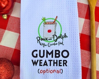 Może przedstawiać: Biały ręcznik kuchenny z czerwoną i zieloną ilustracją garnka z gumbo. Na ręczniku nadrukowany jest tekst "Roux-Dolph The Gumbo Pot" i "GUMBO WEATHER (optional)".