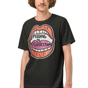 May include: Black t-shirt with a cartoon lip graphic in red and pink, and the words "FEEDING the ALGORITHM". The design has a white outline. The shirt is worn by a person.
