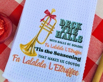Może przedstawiać: Biały ręcznik kuchenny z pozłacaną trąbką i tekstem "Deck the Halls with balls of boudin Fa Lalalala L'Etouffee 'Tis the Seasoning that makes us couyon Fa Lalalala L'Etouffee"