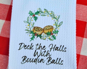 Może przedstawiać: Biały ręcznik kuchenny z zielonym wieńcem z ostrokrzewu i dwoma żółtymi kulkami boudin. Tekst "Deck the Halls With Boudin Balls" jest napisany czarnym kursywą.