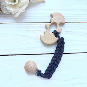 May include: A wooden baby teether in a light brown color, featuring a unique shape with rounded edges. It is attached to a navy blue braided strap with a wooden clip. The teether is designed for babies and toddlers.