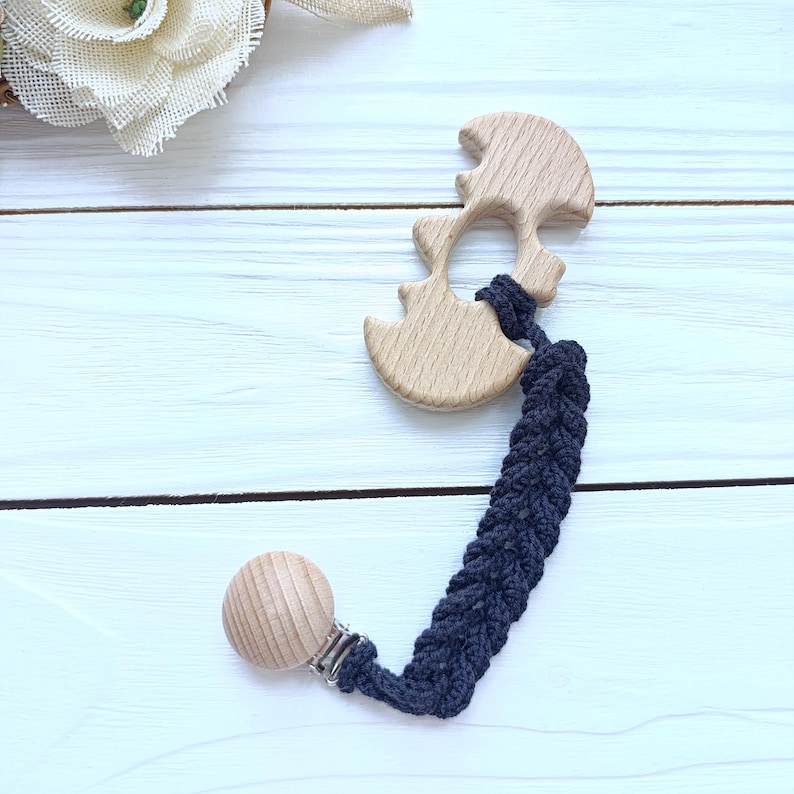 May include: A wooden baby teether in a light brown color, featuring a unique shape with rounded edges. It is attached to a navy blue braided strap with a wooden clip. The teether is designed for babies and toddlers.