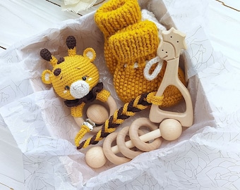 May include: A gift box filled with baby items, including a knitted yellow and white baby bootie, a crocheted giraffe rattle, and a wooden teether shaped like a giraffe. The items are nestled in white tissue paper.