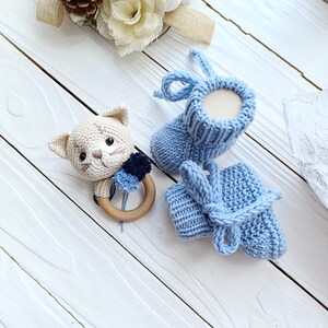 Op de afbeelding: Een paar gebreide babyschoentjes in lichtblauw. E&eacute;n schoentje heeft een strik en het andere een simpele boord. Een gehaakte kattenrammelaar met een houten ring is ook inbegrepen.