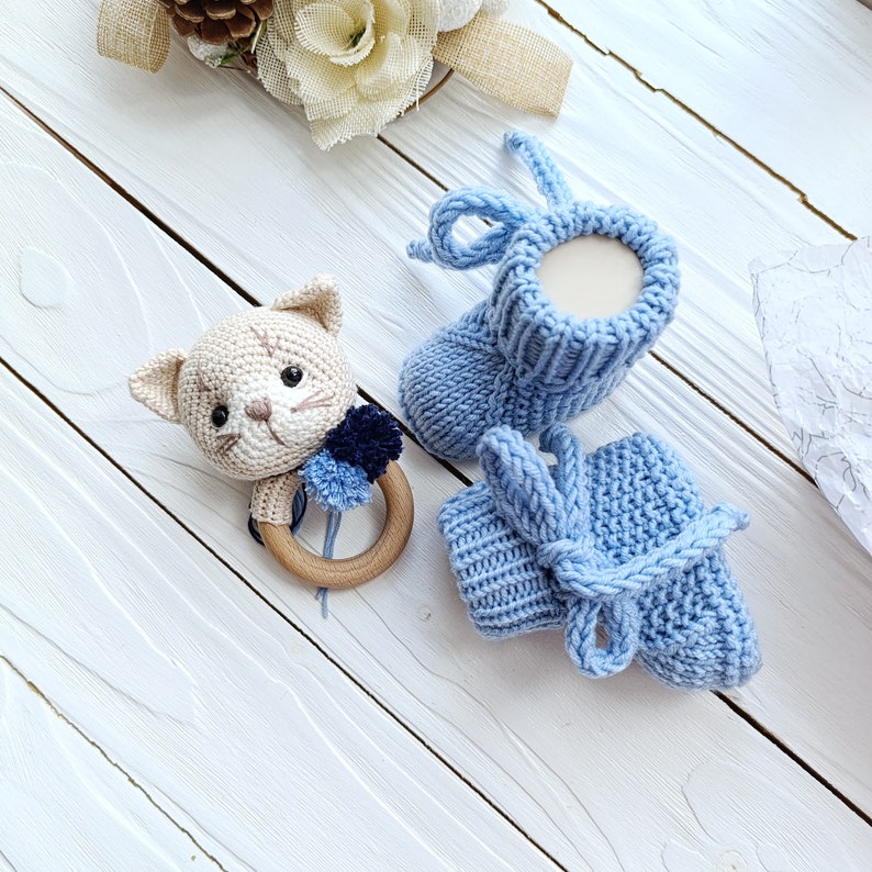 Op de afbeelding: Een paar gebreide babyschoentjes in lichtblauw. E&eacute;n schoentje heeft een strik en het andere een simpele boord. Een gehaakte kattenrammelaar met een houten ring is ook inbegrepen.