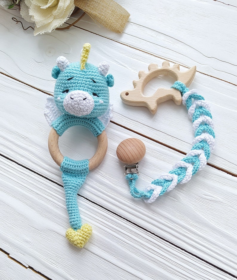 May include: A blue crocheted baby rattle with a wooden ring and a yellow heart. The rattle is attached to a braided cord with a wooden teether and a clip.