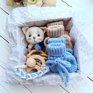 Op de afbeelding: Een cadeau mand gevuld met een gehaakt katten speelgoed, een houten bijtring en twee paar gebreide babyschoentjes. De mand is bekleed met wit tissuepapier.