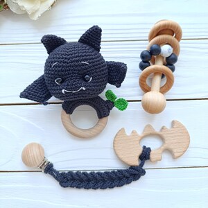 May include: A collection of baby toys, including a crocheted black bat teether with a wooden ring, a wooden rattle with black and white beads, and a wooden bat-shaped teether with a dark blue pacifier clip.