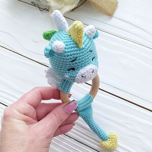 May include: A crocheted blue and white unicorn rattle with a yellow horn and a wooden ring. The rattle has a long tail with a yellow tip.