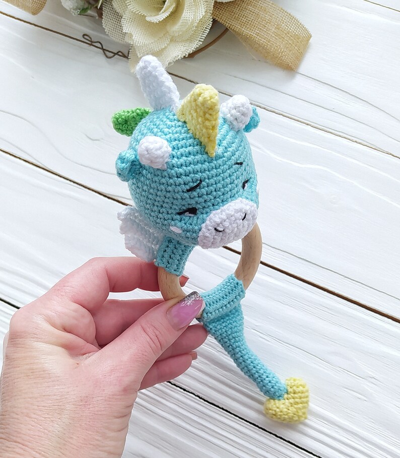 May include: A crocheted blue and white unicorn rattle with a yellow horn and a wooden ring. The rattle has a long tail with a yellow tip.