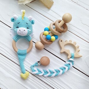 May include: A set of baby toys including a blue crocheted ring with a cute animal head, a wooden rattle with colorful beads, and a wooden teether shaped like a dinosaur. The toys are connected by a braided blue and white strap with a wooden clip.