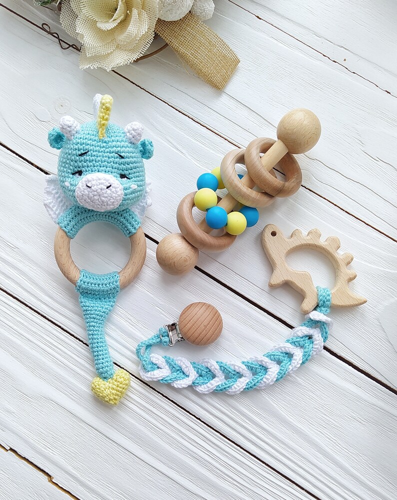 May include: A set of baby toys including a blue crocheted ring with a cute animal head, a wooden rattle with colorful beads, and a wooden teether shaped like a dinosaur. The toys are connected by a braided blue and white strap with a wooden clip.