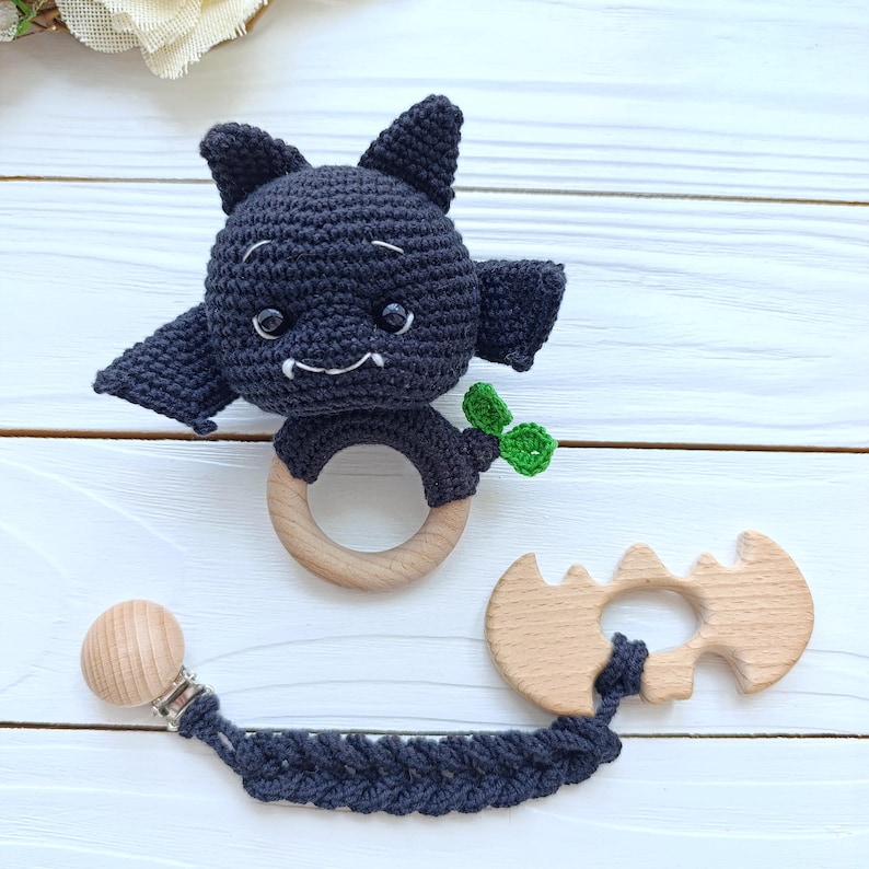 May include: A handmade crochet bat-shaped baby teether in dark gray, with a wooden ring and a small green leaf detail. A matching pacifier clip with a wooden bat-shaped charm is also included.