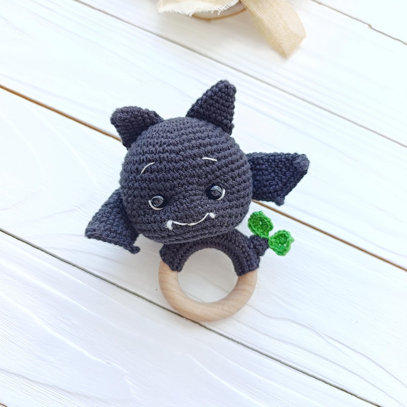 May include: A handmade crochet baby rattle shaped like a dark gray bat with a wooden ring. The bat has small, round eyes, pointed ears, and a smiling mouth. It holds a small green leaf. The rattle is designed for babies.