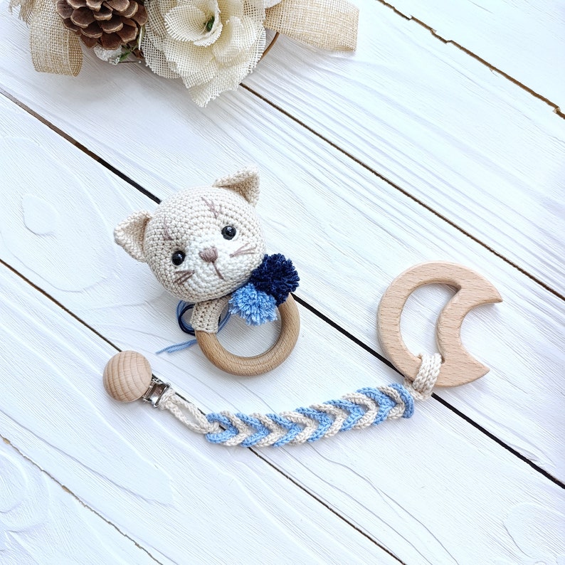 Op de afbeelding: Een gehaakte kattenrammelaar met een houten ring en blauwe pompoenen bevestigd aan een gevlochten band met een houten clip.