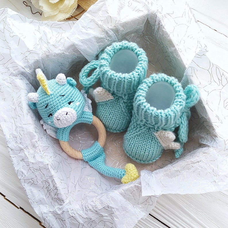 May include: A blue knitted baby bootie set with a crocheted unicorn rattle toy. The booties have white wings and the rattle has a wooden ring.