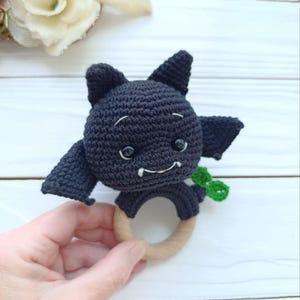 May include: A handmade crochet bat toy in black, featuring small wings, pointy ears, and a smiling face with white details. The bat is attached to a natural wooden ring, perfect for teething. A small green accent is visible.