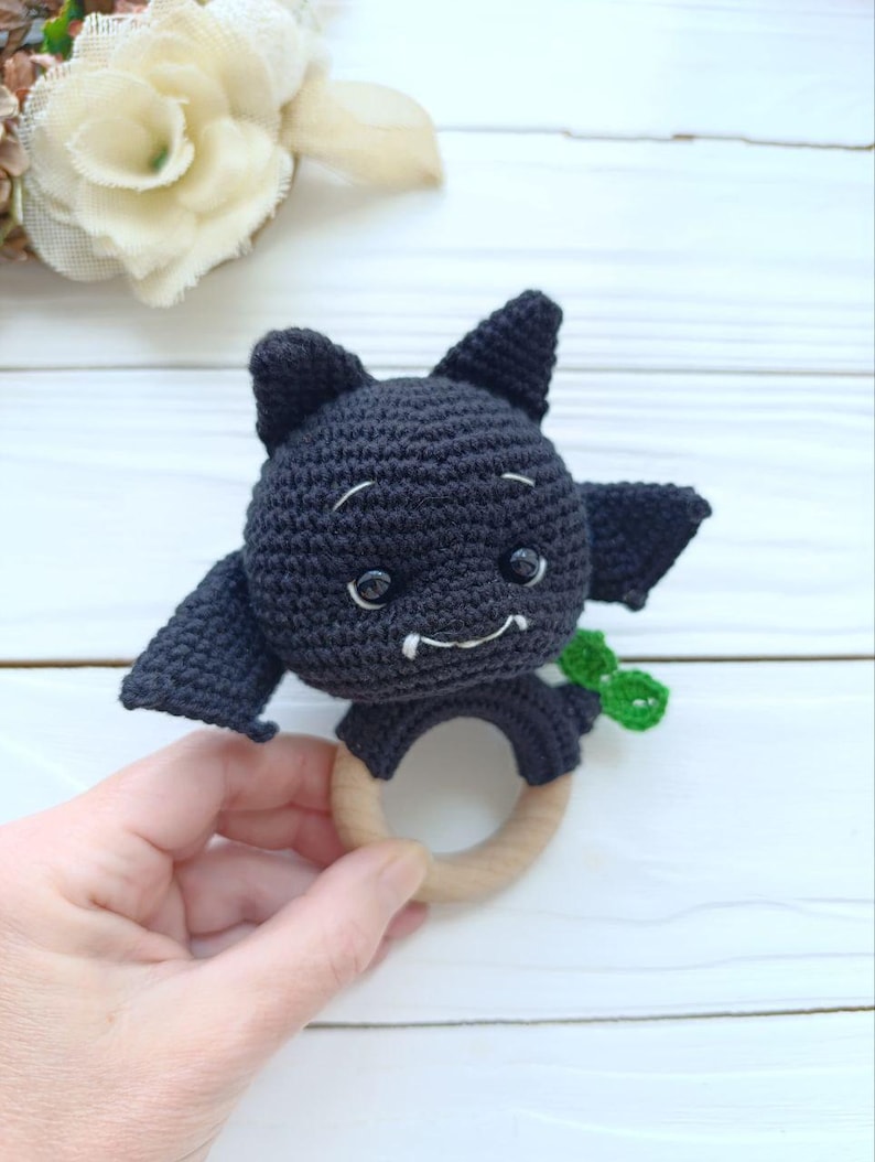 May include: A handmade crochet bat toy in black, featuring small wings, pointy ears, and a smiling face with white details. The bat is attached to a natural wooden ring, perfect for teething. A small green accent is visible.