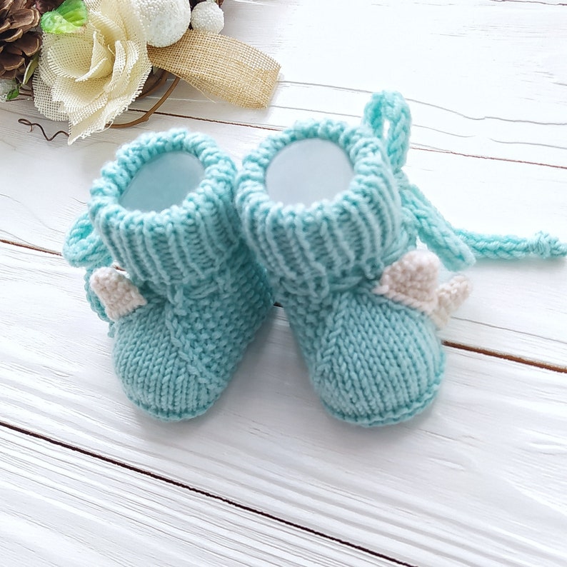 May include: A pair of light blue knitted baby booties with white felt accents. The booties have a simple design and are perfect for a newborn.
