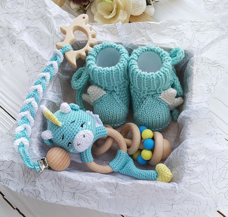 May include: A baby gift set with a blue knitted unicorn rattle, a wooden teether, a blue knitted baby bootie, and a blue knitted baby sock.