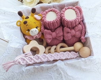 May include: A gift box filled with baby items, including a knitted giraffe toy, a pair of pink knitted booties, a wooden teether, and a pink crocheted strap.