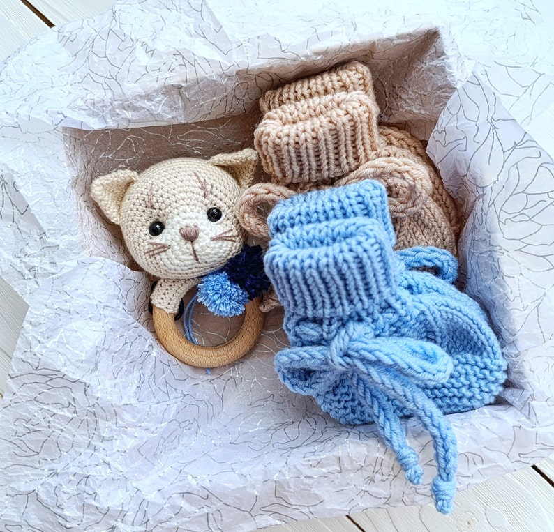 Op de afbeelding: Een cadeaudoos gevuld met gehaakte babyartikelen, waaronder een blauwe gebreide laars, een beige gebreide laars en een gehaakte kattenrammelaar.