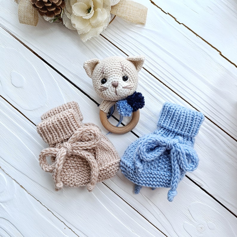 Op de afbeelding: Twee gebreide babyschoentjes, een in beige en een in lichtblauw, met een gehaakt kattenrammelaar speelgoed in het midden. Het rammelaar speelgoed heeft een bruin en wit kattengezicht en een blauwe en witte pompon.