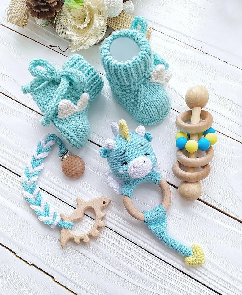 May include: A set of baby toys including a pair of blue knitted booties, a blue knitted unicorn rattle, a wooden teether, and a wooden rattle with blue and yellow beads.