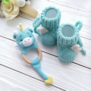 May include: A blue crocheted baby rattle shaped like a dragon with a wooden ring. Two light blue knitted baby booties with white wings.