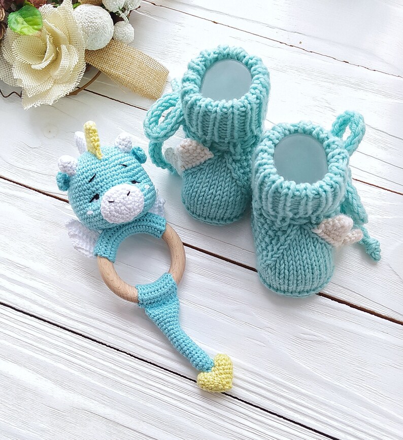 May include: A blue crocheted baby rattle shaped like a dragon with a wooden ring. Two light blue knitted baby booties with white wings.