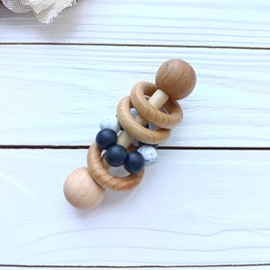 May include: A wooden baby rattle with a light wood finish. The rattle features two wooden spheres at each end, connected by wooden rings and a central section with black and white beads. The design is simple and natural.