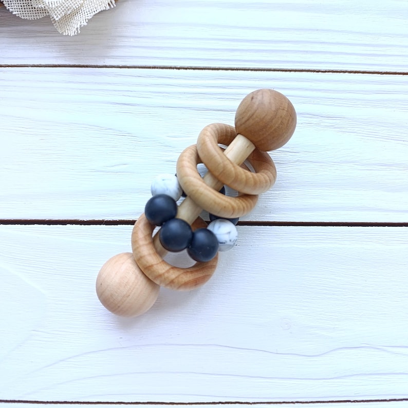 May include: A wooden baby rattle with a light wood finish. The rattle features two wooden spheres at each end, connected by wooden rings and a central section with black and white beads. The design is simple and natural.