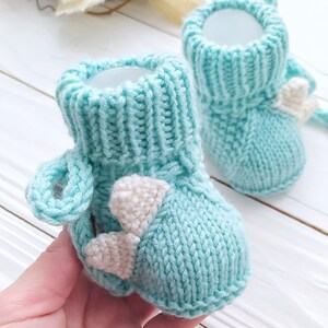 May include: A pair of light blue knitted baby booties with white felt accents in the shape of hearts. The booties have a fold-over cuff and a loop at the top for easy on and off.