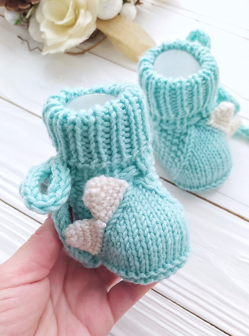 May include: A pair of light blue knitted baby booties with white felt accents in the shape of hearts. The booties have a fold-over cuff and a loop at the top for easy on and off.