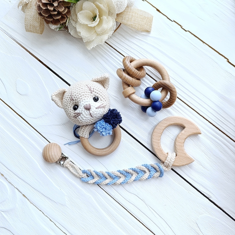 Op de afbeelding: Een set van drie houten baby-bijtjes, waaronder een kattenvormige bijtje met een blauwe pompon, een ring-bijtje met blauwe en witte kralen en een halve maan-bijtje. De bijtjes zijn verbonden door een gevlochten koord met een clip.