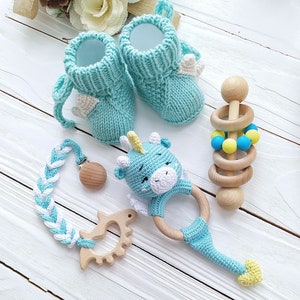May include: A set of baby items including a pair of blue knitted booties with white wings, a blue and white crocheted teething ring with a unicorn head, a wooden teether with a blue and yellow bead ring, and a blue and white braided teething toy with a wooden clip.