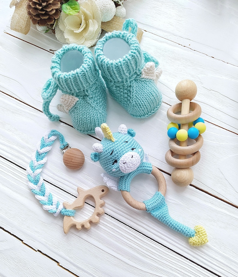 May include: A set of baby items including a pair of blue knitted booties with white wings, a blue and white crocheted teething ring with a unicorn head, a wooden teether with a blue and yellow bead ring, and a blue and white braided teething toy with a wooden clip.