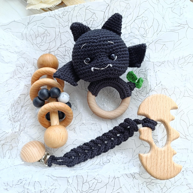 May include: A handmade baby gift set featuring a black crochet bat-shaped rattle with wooden ring, a wooden teether, and a pacifier clip. The set includes wooden beads and a black crochet strap. The bat has small green accents.