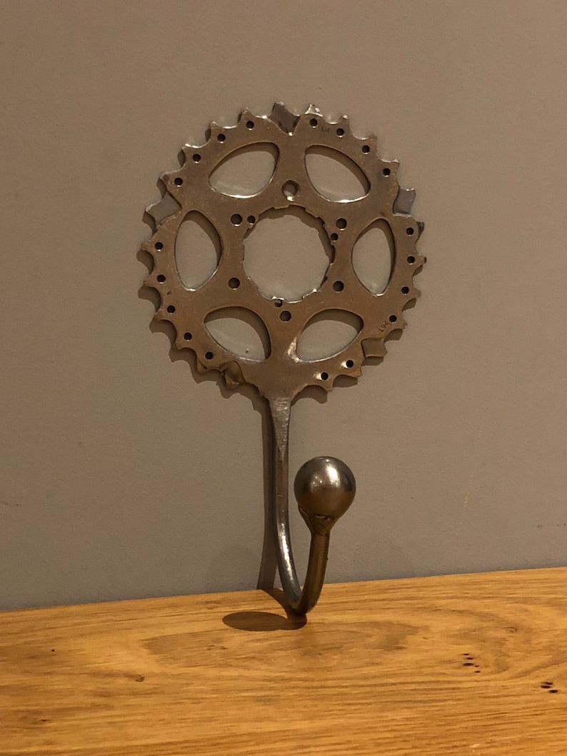 Puede incluir: Un gancho de pared de metal plateado con un dise&ntilde;o de engranaje redondo.