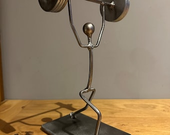 May include: A silver metal sculpture of a person lifting a barbell. The sculpture is made of welded metal rods and is on a rectangular base.